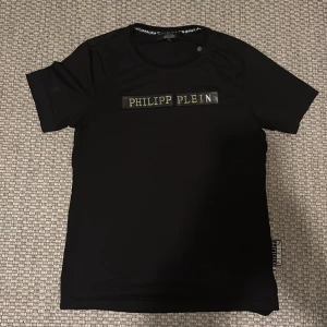  Philipp Plein t-shirt - Svart t-shirt från Philipp Plein med klassisk passform och korta ärmar. T-shirten har ett stilrent tryck med märkesnamnet i guld och silver på bröstet samt en diskret etikett i sidan. PP logga även på nacken. Storlek L men liten i storleken så passar M oxå. 