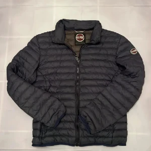 Svart pufferjacka från Colmar - Snygg svart pufferjacka från Colmar med quiltad design och dragkedja framtill. Jackan har hög krage och Colmar-logga på ärmen. Perfekt för kyliga dagar och ger en clean, sportig look.