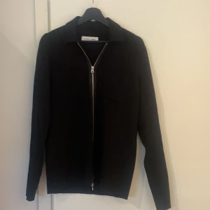 Svart kofta med dragkedja från Samsøe - Stilren svart kofta från Samsøe Samsøe med hel dragkedja framtill och klassisk krage. Tröjan har en bröstficka och är långärmad. Perfekt för lager-på-lager och enkel att matcha med det mesta.