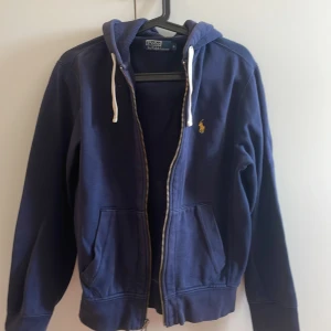 Marinblå hoodie från Polo Ralph Lauren - Snygg marinblå hoodie från Polo Ralph Lauren med dragkedja, huva med vita snören och klassisk gul logga på bröstet. Tröjan har två fickor framtill och är tillverkad i mjuk bomull, perfekt för en chill och stilren look.