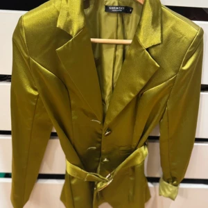 Grön satin kavaj  - Snygg grön kavaj i glansigt satinmaterial    Kavajen har markerad midja med bälte, klassisk krage och knappar framtill. Långärmad och figurnära passform. 