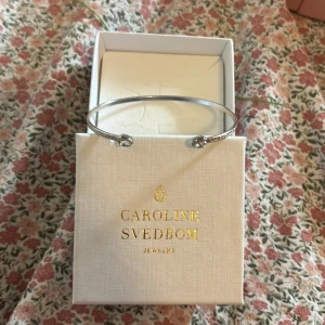 Silverfärgat armband Caroline Svedbom - Stilrent och tunt armband i silverfärg från Caroline Svedbom. Armbandet är öppet med två små kristaller i ändarna som ger en elegant touch. Perfekt att kombinera med andra smycken för en personlig look. Ordinarie pris 700kr