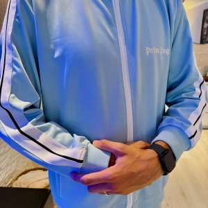 Ljusblå Palm Angels zip hoodie - Säljer en ljusblå hoodie från Palm Angels med vit dragkedja och vita ränder med svarta detaljer längs ärmarna. Tröjan har hög krage, ribbade muddar och logga på bröstet. Perfekt för en sportig och trendig look. Passar bra som ovesrize modell.