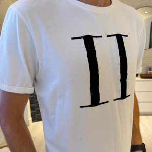 Vit t-shirt med svart tryck från Les Deux - Vit t-shirt från Les Deux med stort svart II-tryck på bröstet. Klassisk rund halsringning och korta ärmar. Skön passform och tillverkad i mjuk bomull, perfekt för en clean och stilren look. Passar perfekt s/m storlek.