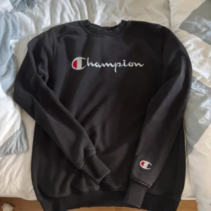 Svart sweatshirt från Champion M - Svart sweatshirt från Champion i storlek medium. Tröjan har klassisk rund hals, ribbade muddar och det ikoniska Champion-loggan broderad på bröstet samt på ärmen. Perfekt för en avslappnad och sportig stil.