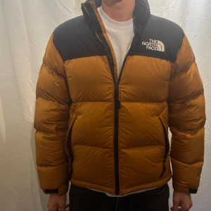 The North Face pufferjacka - Grym pufferjacka från The North Face i svart och brunt. Jackan har hög krage, dragkedja framtill och två sidofickor med dragkedja. Fylld med dun och har en klassisk  passform. Perfekt för kalla dagar och riktigt snygg streetstil. Modellen är 180cm