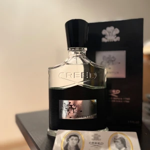 Creed Aventus Eau de Parfum 100ml - Lyxig parfym från Creed, Aventus, i en elegant glasflaska med svart och silverdetaljer. Flaskan har en svart kork och en metallisk etikett med ryttare. Kommer med originalförpackning och Creed-kort. Perfekt för dig som gillar exklusiva dofter.