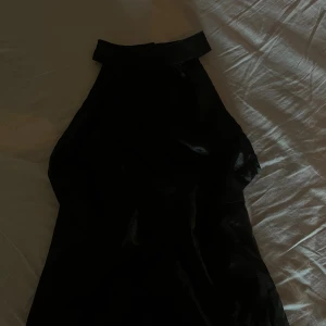 Svart topp halterneck - Snygg svart topp med halterneck och hög krage. Toppen är ärmlös och har en slät, glansig yta som ger en cool vibe. Perfekt för dig som gillar minimalistisk stil och vill sticka ut med en enkel men elegant look.