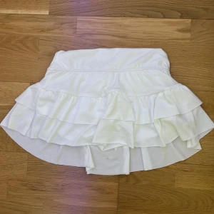 Vit volangkjol med shorts under - Supersöt vit kortkjol med dubbla volanger och inbyggda shorts under för extra komfort. Kjolen har en mjuk och stretchig känsla, perfekt för en sportig och trendig look. Passar perfekt till sneakers och en crop top för en fräsch outfit.