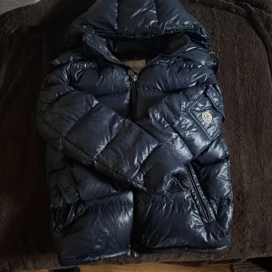 Mörkblå Moncler dunjacka med huva - Snygg mörkblå dunjacka från Moncler med glansig finish och vadderad design. Jackan har huva, dragkedja framtill och två dragkedjeförsedda fickor. Klassisk Moncler-logga på ärmen och insida i kontrasterande brun nyans. Perfekt för kalla dagar sjukt varm jacka säljer billigt pga slit och trasig inne ficka 