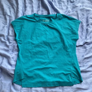 Turkos t-shirt från New Apparel XS - Turkos t-shirt från New Apparel i storlek XS. Modellen är enkel med rund halsringning och korta ärmar. Materialet är mjukt och lätt, perfekt för en avslappnad stil. Passformen är normal och t-shirten har diskreta ränder i tyget.
