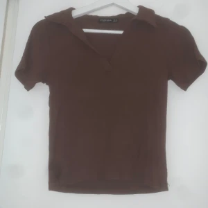 Brun ribbad topp  - Brun ribbad topp från Stradivarius med korta ärmar och krage. Toppen har en v-ringad öppning framtill och är i ett stretchigt material som sitter snyggt på kroppen.