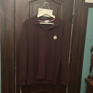 Vinröd långärmad pikétröja Moncler - Vinröd långärmad pikétröja från Moncler med klassisk krage och diskret logotyp på bröstet. Tröjan är i mjuk bomull och har en stilren look som passar perfekt till jeans eller chinos. Snygg och enkel att matcha med andra plagg. Storlek M-S