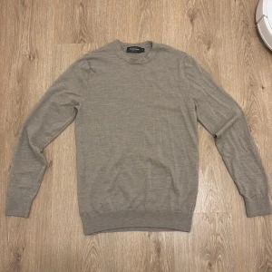 Beige merinoullströja Dressmann - En stilren beige tröja från Dressmann i mjuk merinoull. Klassisk rund halsringning och långärmad modell med ribbade muddar vid ärmslut och nederkant. Perfekt för lager-på-lager och enkel att matcha med olika outfits.