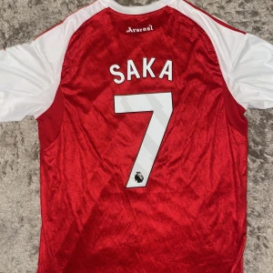 Arsenal Saka 7 matchtröja Adidas L - Riktigt snygg Arsenal fotbollströja med Saka 7 på ryggen. Röd bas med vita ärmar och detaljer, broderat klubbmärke och Adidas-logga. Tillverkad i lätt och ventilerande polyester med diskret mönster i tyget. Emirates Fly Better tryck på bröstet.