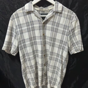 Rutig kortärmad skjorta från Primark - Kortärmad skjorta från Primark . Skjortan är vit med grå och beige rutigt mönster, har klassisk krage och knäppning framtill. Tillverkad i ett strukturerat styg som känns både luftigt och stilrent. Tröjan står att det är XS men den passar S. Den är köpt i Spanien och där har de väldigt skumma storlekar. Säljer den för att jag har en liknande som jag använder mer. Storlek S (xs) skick 7/10.