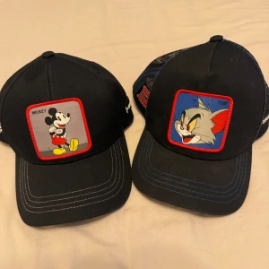 Svarta kepsar med Mickey och Tom - Två svarta kepsar med broderade patchar framtill, en med Mickey Mouse och en med Tom från Tom & Jerry. Kepsarna har böjd skärm, kontrastsömmar och mesh på sidorna. Perfekt för dig som gillar klassiska tecknade figurer.