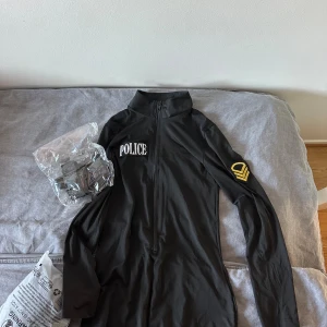 Svart långärmad police-dräkt - Svart långärmad dräkt med dragkedja framtill och vit 'POLICE'-text på bröstet. På ena ärmen finns ett gult emblem. Tröjan har en hög krage och är gjord i ett syntetmaterial som känns smidigt och lätt. Perfekt för temafest eller halloween, tillkommer band man kan ha runt benen och midjan. SÄLJER 2 st liknande