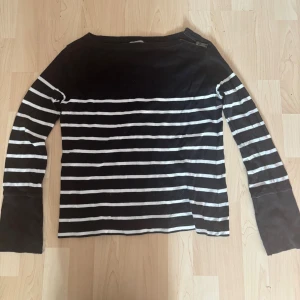 Randig svartvit tröja från Hunkydory - Svart och vit randig långärmad tröja från Hunkydory. Tröjan har en cool dragkedjedetalj vid axeln och breda muddar vid ärmsluten. Tillverkad i mjuk bomull och har en avslappnad passform. Perfekt för en stilren och casual look.