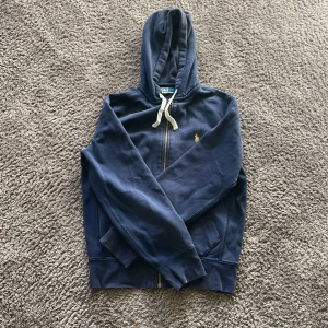 Blå hoodie från Polo Ralph Lauren - Mörkblå hoodie från Polo Ralph Lauren med dragkedja, huva och vita snören. Klassisk gul broderad logga på bröstet och två fickor framtill. Tillverkad i mjuk bomull, perfekt för chill dagar. Snygg och enkel att matcha med jeans eller joggers. Skicket skulle jag anse som att vara väldigt bra för den har inga defekter priset är inte hugget i stenen 