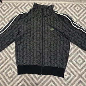 Svart mönstrad vindjacka från Lacoste - Snygg svart vindjacka från Lacoste med geometriskt mönster och klassiska vita ränder längs ärmarna. Jackan har dragkedja framtill, hög krage och ribbade muddar. Ikoniska gröna krokodilloggan på bröstet ger extra stilpoäng.