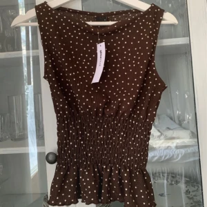 Brun prickig blus från Gina Tricot - Supersöt brun blus med vita prickar från Gina Tricot. Modellen är ärmlös med smockad midja och volangkant nedtill som ger en snygg siluett. Materialet är mjukt och lätt, perfekt för varmare dagar. Prickigt mönster gör den extra charmig! Storlek XS