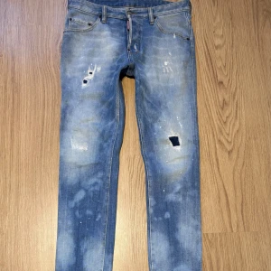 Dsquared2 Jeans Blå 46 - Snygga Dsquared2 Jeans i blå tvätt med slitningar, hål och färgstänk för en riktigt edgy look. Klassisk femficksmodell med Dsquared2-logga vid gylfen och bakficka. Tillverkade i Italien av bomull. Perfekta för dig som gillar streetstyle. ÄKTA