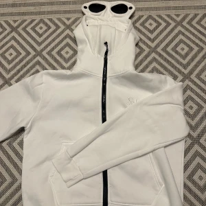 Vit C.P. Company hoodie med goggles - Säljer en vit hoodie från C.P. Company med ikoniska goggles i huvan. Tröjan har hel dragkedja framtill, fickor och broderad logga på bröstet. Perfekt för dig som vill sticka ut med streetwear-vibe och coola detaljer.