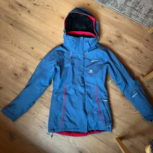 Blåskidjacka från Salomon - Fin skidjacka från Salomon använd endast en vecka, inga tecken på användning och som i nyskick! Jackan har en figurnära form med snölås, flertal fick som innerficka och för liftkort, avtagbar luva och foder på insidan. Nypris 2500!