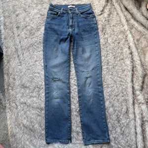 Blå raka mid waist jeans med slitningar  - Blå jeans med knapp och dragkedja. Stretchigt material. Revor på båda knäna varav två är lagade. Rak modell. Jättesköna.  Innerbenslängd ca 79 cm Midja tvärs över ca 36 cm