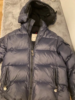 Mörkblå dunjacka från Moncler - Snygg mörkblå dunjacka från Moncler med huva och två dragkedjeförsedda fickor. Jackan har quiltad design, hög krage och klassisk Moncler-logga på ärmen. Perfekt för kalla dagar och riktigt stilren look.