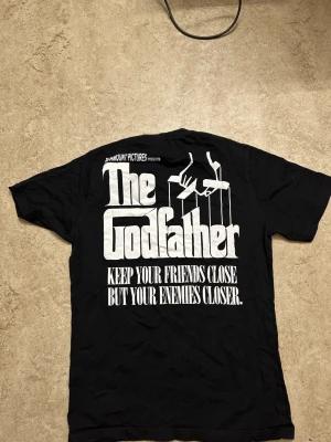 The Godfather svart t-shirt - Svart t-shirt med stort The Godfather-tryck på ryggen och ikoniskt motiv från filmen på framsidan. Klassisk passform med korta ärmar och rund hals. Perfekt för filmälskare som vill sticka ut med en cool och tidlös design.