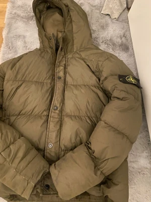 Olivgrön pufferjacka från Stone Island - Säljer en olivgrön pufferjacka från Stone Island med ikonisk patch på ärmen. Jackan har huva, dragkedja och knappar framtill samt quiltad design. Perfekt för kalla dagar och riktigt snygg streetwear vibe. Storlek 152 till 160.