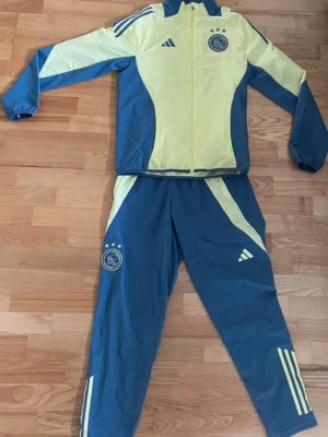 Ajax träningsset Adidas blå/gul - Ajax träningsset från Adidas i blått och ljusgult. Setet består av en jacka med dragkedja och matchande byxor, båda med Ajax-logga och Adidas-tryck. Snygga kontrastpaneler och klassiska ränder på benen. Perfekt för dig som gillar fotboll och vill sticka ut på planen.