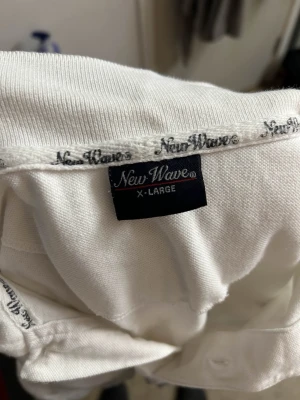 Vit pikétröja från New Wave - Klassisk vit pikétröja från New Wave i storlek XL. Tröjan har korta ärmar, krage och knappar framtill samt ett broderat märke med tre stjärnor på bröstet. Perfekt till en clean och enkel stil.
