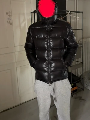 Svart pufferjacka från Moncler - Snygg svart pufferjacka från Moncler med glansig finish och hög krage. Jackan har huva, dragkedja framtill och två sidofickor. Klassisk Moncler-logga på ärmen och quiltad design som ger en riktigt fet vinterlook.