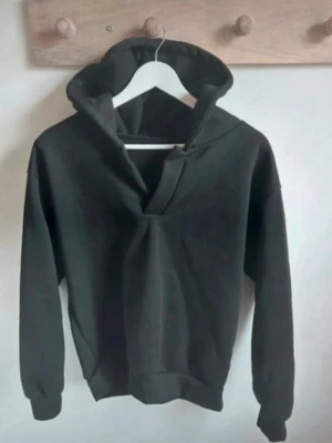 Ny, Svart hoodie med v-ringning - Säljer en svart hoodie med unik v-ringad hals och bred huva. Hoodien har långa ärmar och ribbade muddar vid ärmslut och nederkant. Tillverkad i mjukt sweatshirtmaterial som känns skönt mot huden. Perfekt för en avslappnad och trendig look.