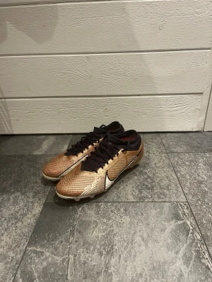 Nike mercurial fotbollsskor - Snygga fotbollsskor från Nike i guld med svarta detaljer och vit swoosh. Skorna har snörning och en låg profil som ger bra bollkontroll. Perfekta för dig som vill sticka ut på planen med stilrena och sportiga skor. Nypris ca 2500kr☺️