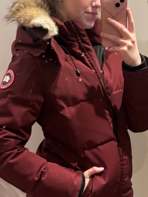 Vinröd dunjacka från Canada Goose - Snygg vinröd dunjacka från Canada Goose med pälskantad huva och klassisk logga på ärmen. Jackan har dragkedja framtill, hög krage och justerbar huva. Perfekt för kalla vinterdagar och riktigt varm.