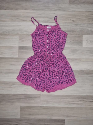 Rosa jumpsuit med bokstavsmönster - Snygg rosa jumpsuit med svarta bokstäver och vita detaljer. Smala axelband, knappar framtill och resår i midjan för skön passform. Perfekt för dig som vill ha en lekfull och trendig stil. Från Bench i stl 140.