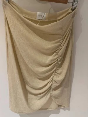 Glittrig beige kjol från Vila storlek S - Supersnygg beige kortkjol från Vila med glittrigt tyg och draperad effekt framtill. Kjolen har en tight passform och är perfekt för dig som vill sticka ut lite extra. Materialet känns mjukt och följsamt mot kroppen. Längd 56cm