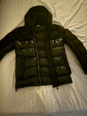 Svart pufferjacka från KWD - Snygg svart pufferjacka från KWD med glansiga partier och broderad logga på bröstet. Jackan har huva, dragkedja framtill och quiltad design som ger en modern look. Perfekt för kalla dagar när du vill hålla stilen.