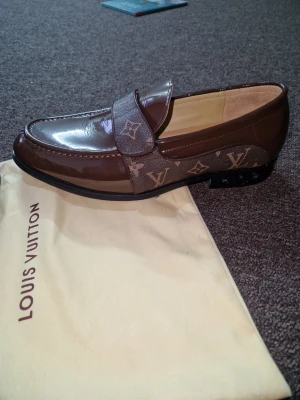 Bruna loafers från Louis Vuitton - Snygga bruna loafers från Louis Vuitton i glansigt skinn med klassiskt LV-monogram på bandet och sidan. Skorna har rund tå och platt sula, perfekt för dig som vill ha en exklusiv touch till din outfit.