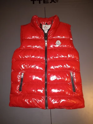 Röd glansig dunväst från Moncler - Säljer en röd Moncler dunväst i klassisk glänsande modell, med originalpatch, tvättetikett och Moncler-detaljer (se bilder). Väldigt snygg färg och perfekt både höst, vinter och vår 🌟 Skick: Bra använt skick, inga hål eller större defekter. Normal användning. Stängs med dragkedja framtill och två fickor med dragkedja. Den är i stolek xs men sitter som s. Perfekt om du vill ha en stilren och tidlös Moncler-väst 🔥 Kan gå ner i pris vid snabb affär 