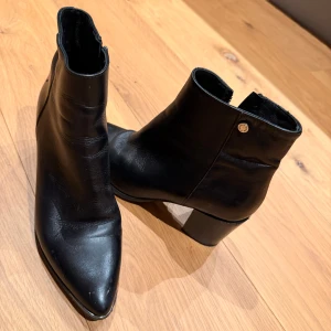 Svarta stövletter med klack - Snygga svarta stövletter i skinn med spetsig tå och bred klack. Klassisk modell med dragkedja på insidan och diskret metallknapp som detalj. Perfekta för att lyfta din outfit med en stilren touch.