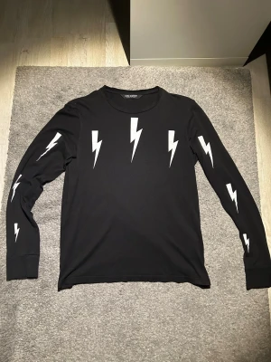 Svart långärmad tröja Neil Barrett - Cool svart långärmad tröja från Neil Barrett med vita blixttryck på bröstet och ärmarna. Tröjan har rund halsringning och är gjord i mjukt material som känns skönt mot huden. Perfekt för dig som gillar streetwear och grafiska detaljer. Passar dig som är runt 170-175🙌🙌
