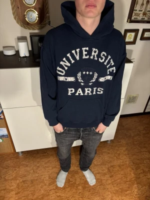 Mörkblå hoodie Université Paris - Mörkblå hoodie med stor vit text 'UNIVERSITÉ PARIS' och tryckt emblem på bröstet. Klassisk känguruficka framtill och stor huva. Skön och avslappnad passform, perfekt till jeans eller joggers. Materialet känns mjukt och bekvämt.
