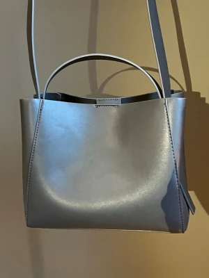Grå axelväska i skinn - Stilren grå axelväska i slätt skinn med minimalistisk design. Väskan har både ett kort handtag och en lång axelrem, vilket gör den enkel att bära på olika sätt. Perfekt för dig som gillar enkel och modern stil. Köpt från Gina Tricot förra året, använts fåtal gånger 🩶