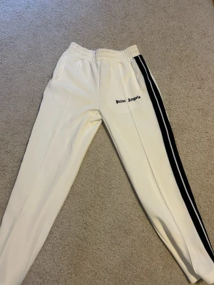 Vita Palm Angels mjukisbyxor med svart stripe - Palm Angels vita mjukisbyxor med svart sidostripes och logga på vänster ben. Byxorna har elastisk midja och är tillverkade i 100% polyester för en sportig och bekväm känsla. Perfekta för en avslappnad och trendig streetwear-look.