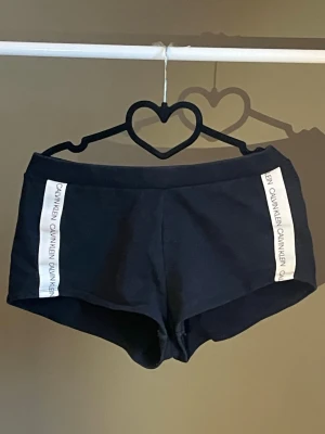 Svarta shorts från Calvin Klein - Svarta shorts från Calvin Klein med vita logoband längs sidorna. De har en tight passform och är tillverkade i mjukt bomullsmaterial. Perfekta för en sportig och avslappnad stil. Köpta från Calvin Klein, använts några gånger🖤🤍